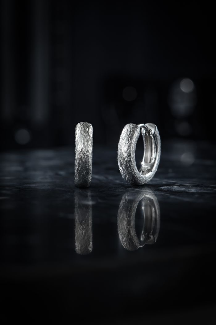 Close-up of textured silver hoop earrings with a dramatic shadow on a reflective surface.
